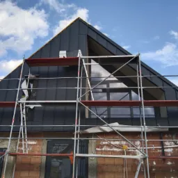 Montaż czarnego, metalowego pokrycia dachu na nowoczesnym domu jednorodzinnym, widoczne rusztowanie i niedokończona elewacja z cegły.