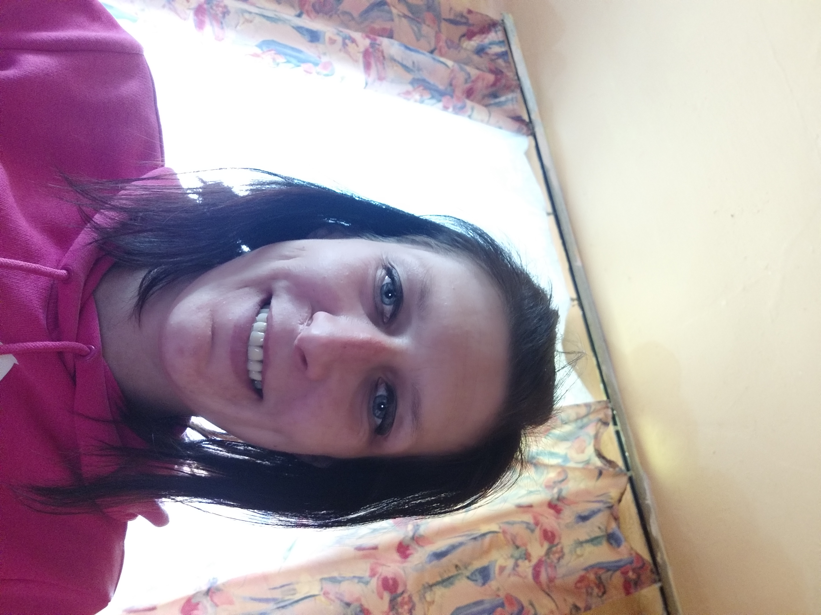 Uśmiechnięta kobieta w różowej bluzie z kapturem, selfie wewnątrz pomieszczenia z zasłonami w tle.