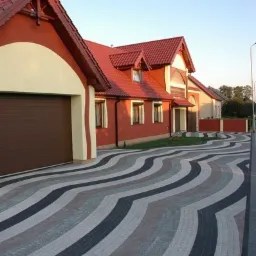 Nowoczesny podjazd z kostki brukowej ułożonej we wzór fal przed domem z czerwoną dachówką i brązową bramą garażową.