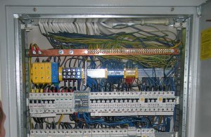 Otwarta skrzynka rozdzielcza z widocznymi bezpiecznikami, przewodami w różnych kolorach i listwą zaciskową z miedzianą szyną uziemiającą.