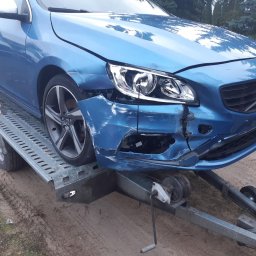 Usługi Blacharsko-Lakiernicze oraz motoryzacyjne - Uszkodzony, błękitny samochód marki Volvo na lawecie, z widocznymi pęknięciami i zarysowaniami na przednim zderzaku i błotniku, z ręką wskazującą na uszkodzenia.