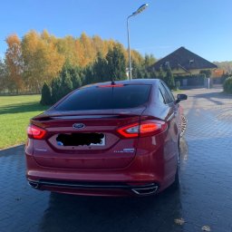 Usługi Blacharsko-Lakiernicze oraz motoryzacyjne - Czerwony Ford Fusion Titanium z widocznymi tylnymi światłami i elementami chromowanymi, zaparkowany na mokrym bruku z jesiennym tłem drzew i domu.