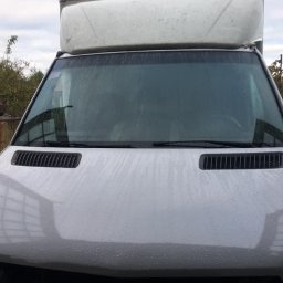 Usługi Blacharsko-Lakiernicze oraz motoryzacyjne - Przednia część brudnego, białego samochodu dostawczego z widocznymi kroplami wody na masce i przedniej szybie, na tle szarego nieba i zabudowań.