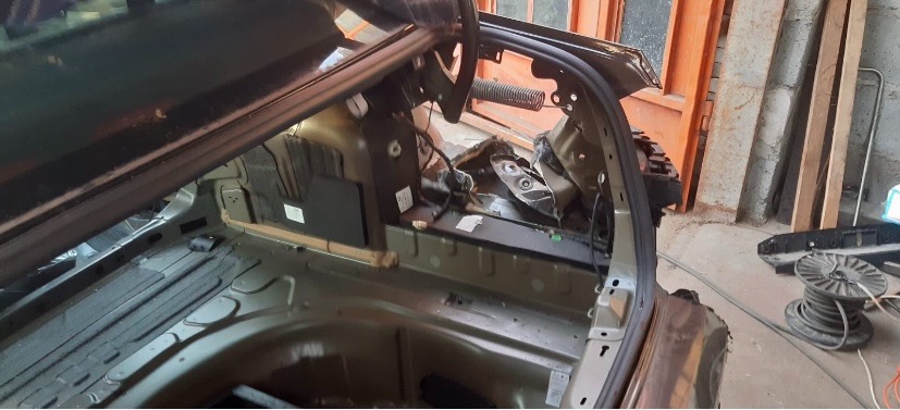 Wnętrze bagażnika samochodu po wypadku, widoczne uszkodzone elementy karoserii i instalacji elektrycznej, w tle fragment warsztatu.