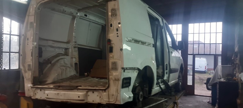 Biały samochód dostawczy na podnośniku w warsztacie, z otwartymi drzwiami i częściowo zdemontowanym tyłem, przygotowywany do naprawy blacharskiej.