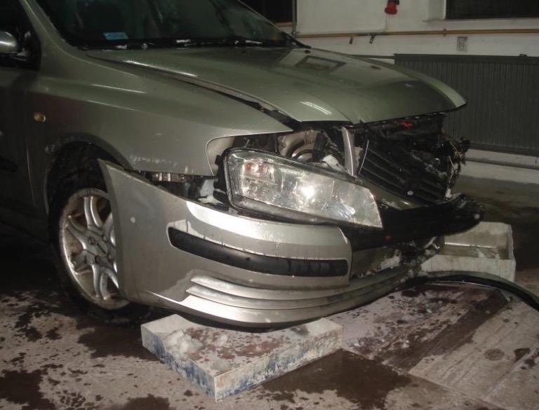 Uszkodzony przód srebrnego samochodu osobowego, z widocznymi zniszczeniami reflektora, zderzaka i karoserii; auto podparte na betonowych blokach w warsztacie.