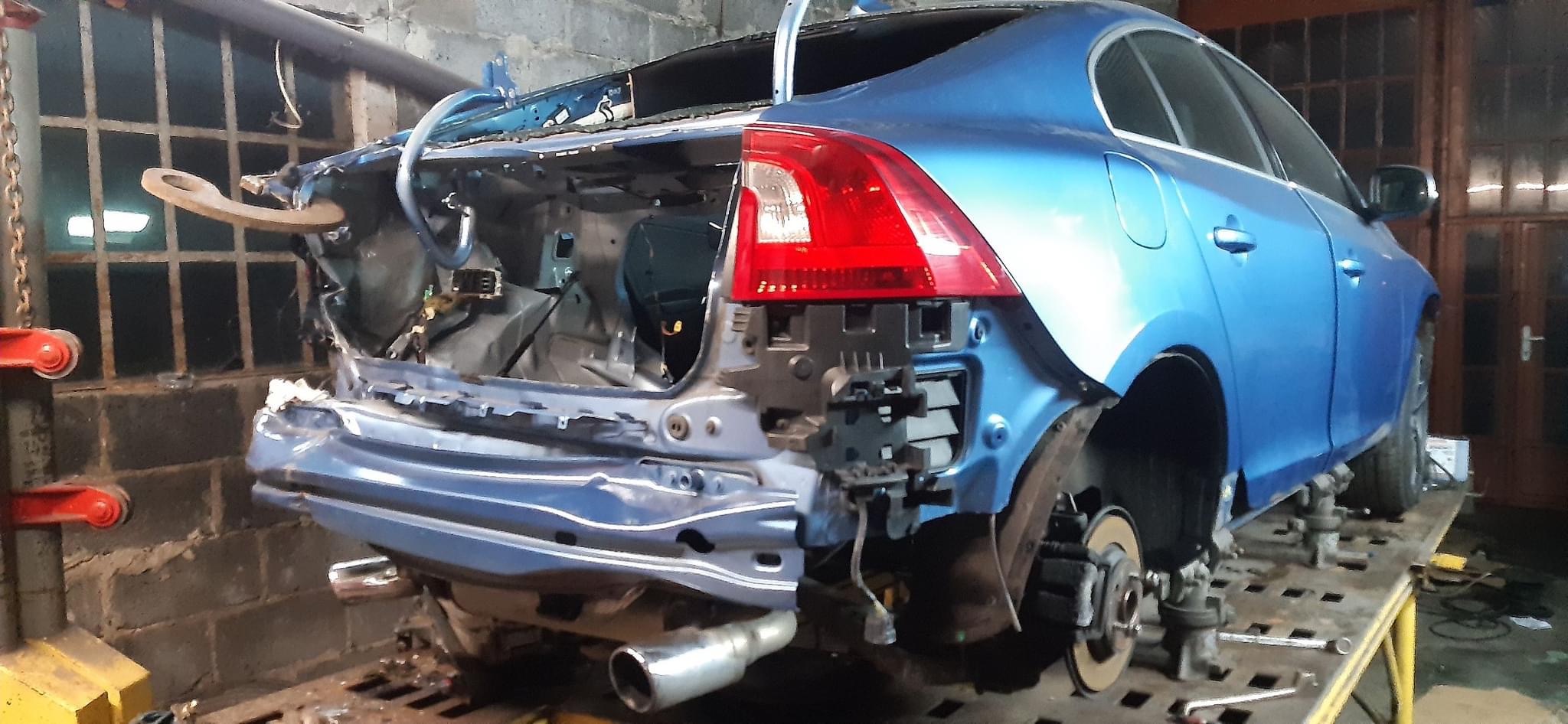Niebieski samochód po wypadku, z uszkodzonym tyłem, w trakcie naprawy blacharskiej w warsztacie, widoczne narzędzia i elementy karoserii.