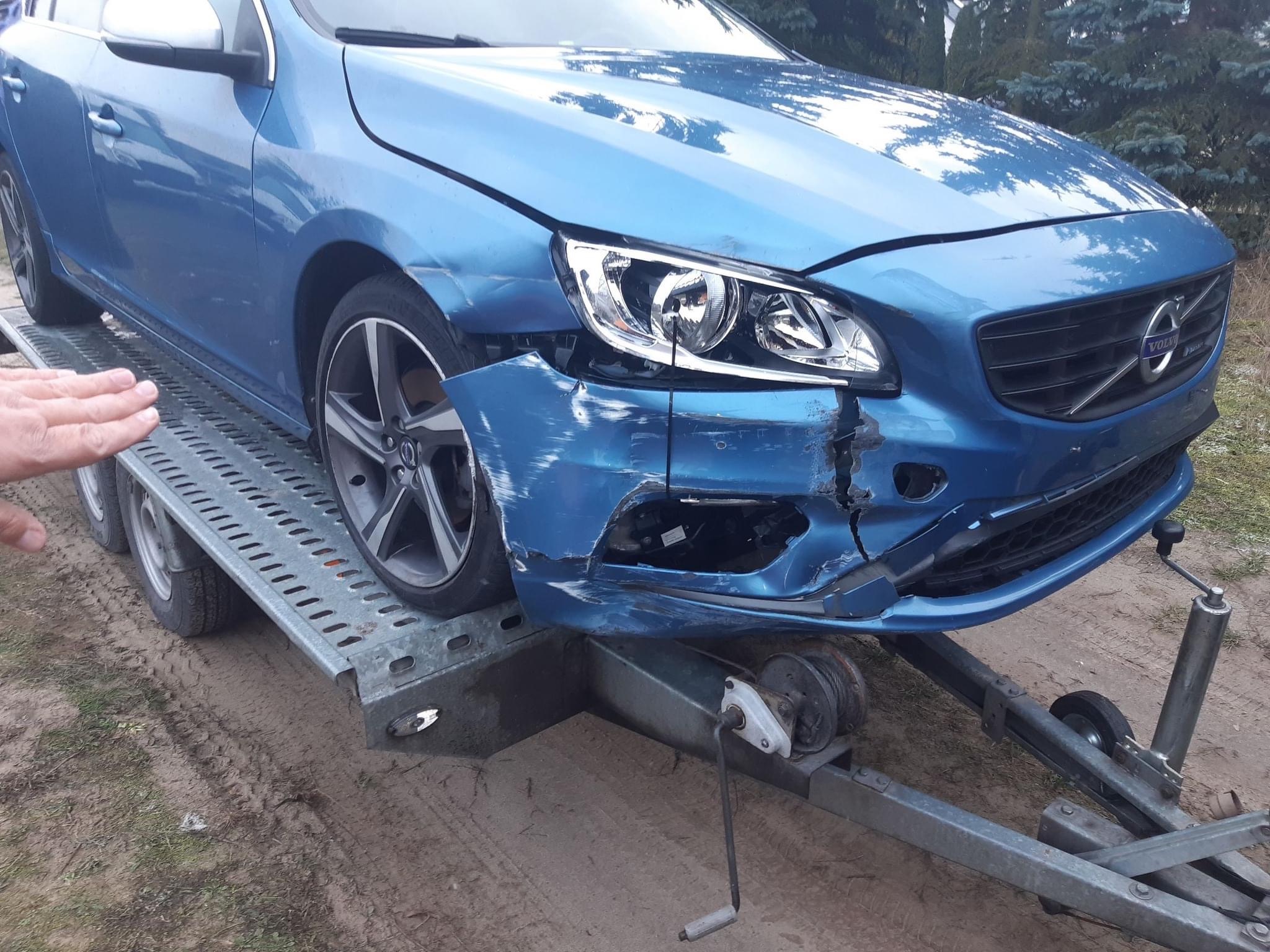 Uszkodzony, błękitny samochód marki Volvo na lawecie, z widocznymi pęknięciami i zarysowaniami na przednim zderzaku i błotniku, z ręką wskazującą na uszkodzenia.