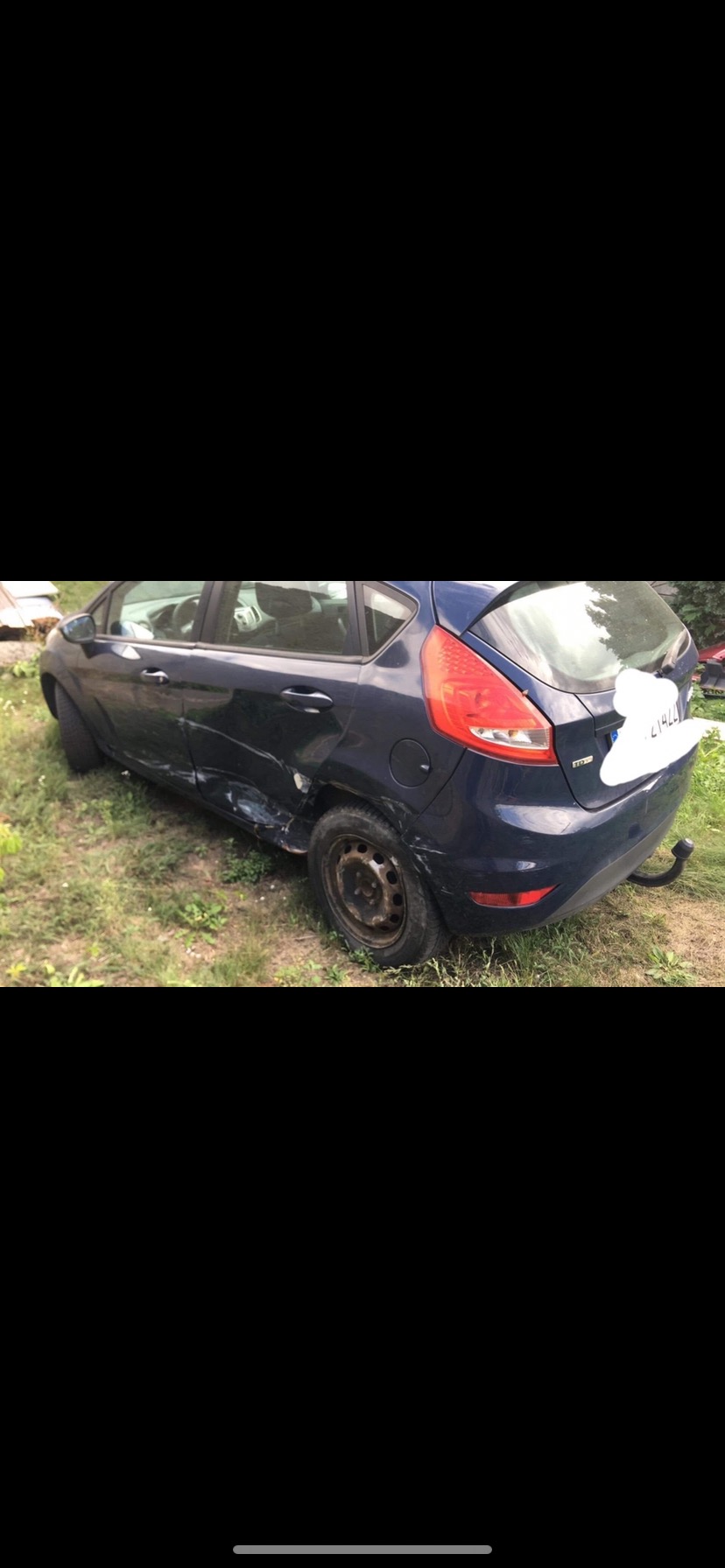 Uszkodzony, granatowy Ford Fiesta stoi na trawie, widoczne wgniecenia na tylnych drzwiach i błotniku, brak kołpaka na tylnym kole.