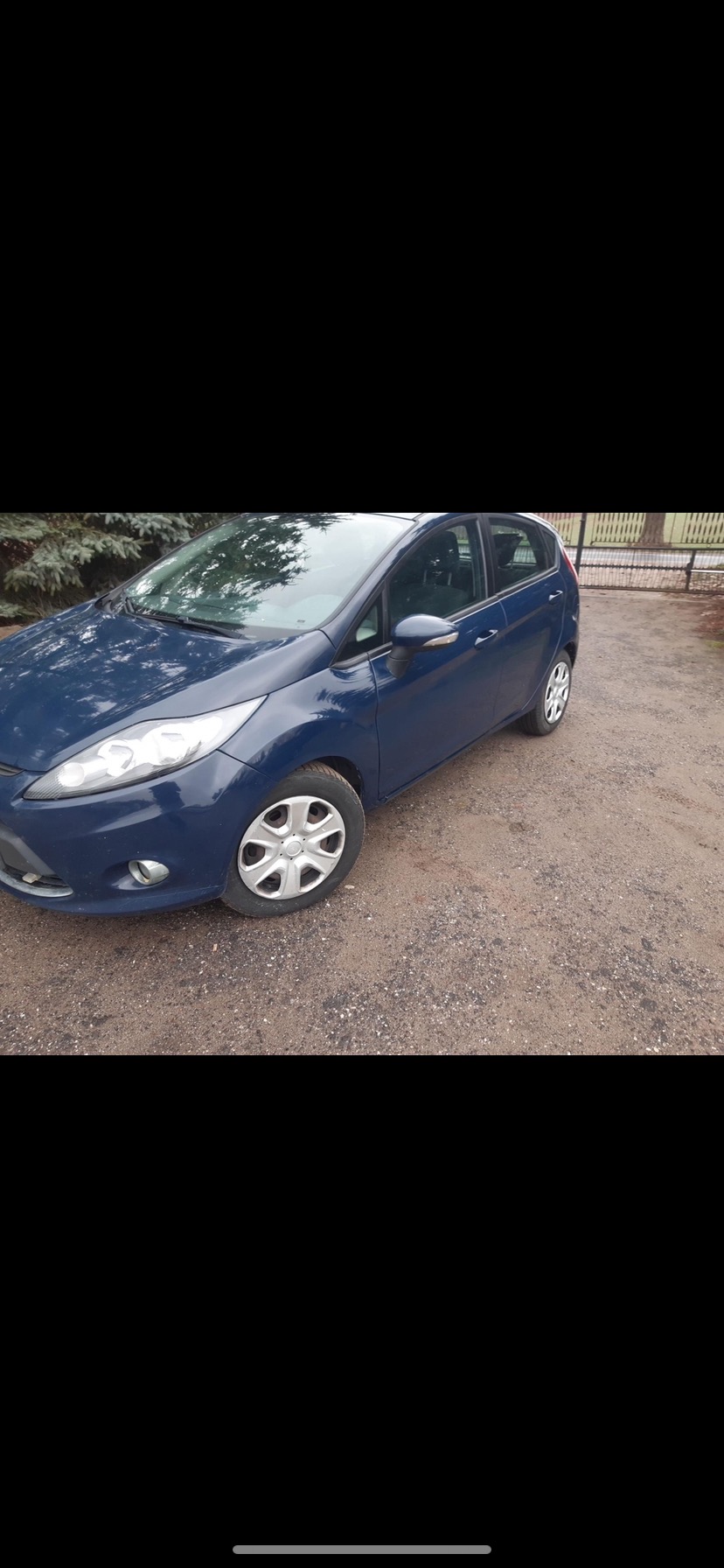 Niebieski Ford Fiesta zaparkowany na żwirowej drodze, widoczne przednie światła i felgi z kołpakami.