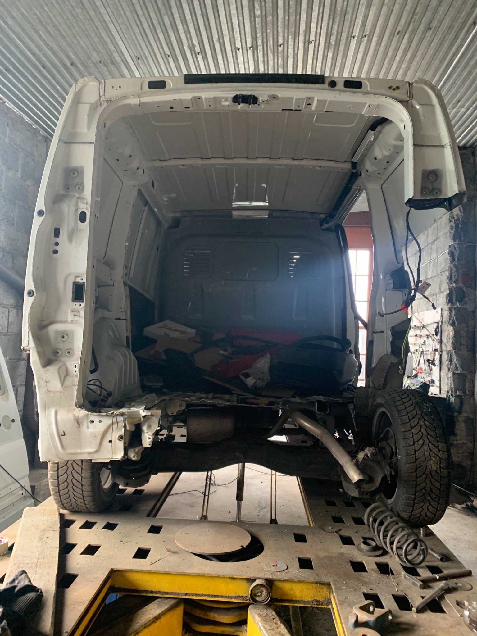 Wnętrze zdekompletowanego, białego samochodu dostawczego na podnośniku warsztatowym. Widoczne uszkodzenia karoserii, elementy zawieszenia i fragmenty instalacji elektrycznej. Tło stanowi ściana...