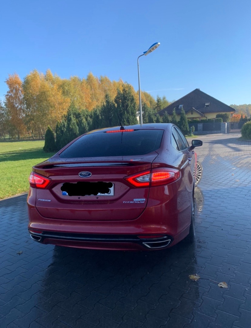 Czerwony Ford Fusion Titanium z widocznymi tylnymi światłami i elementami chromowanymi, zaparkowany na mokrym bruku z jesiennym tłem drzew i domu.