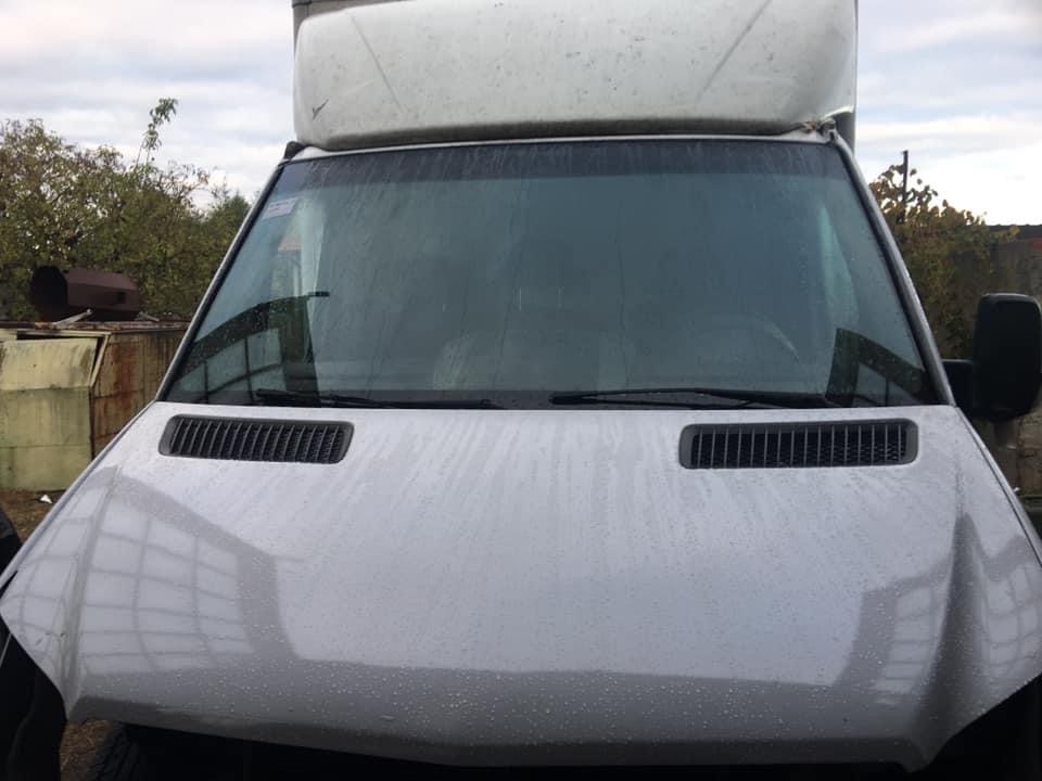 Przednia część brudnego, białego samochodu dostawczego z widocznymi kroplami wody na masce i przedniej szybie, na tle szarego nieba i zabudowań.