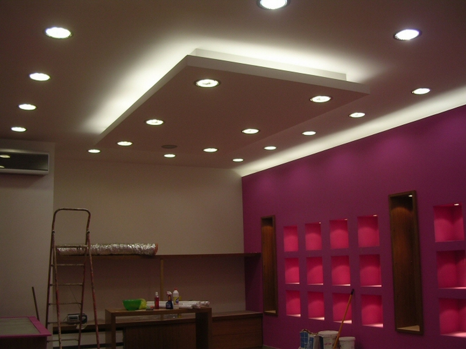 Wnętrze w trakcie wykończenia z podwieszanym sufitem z oświetleniem punktowym i dekoracyjną ścianą w kolorze fuksji z wnękami, widoczna drabina i narzędzia.