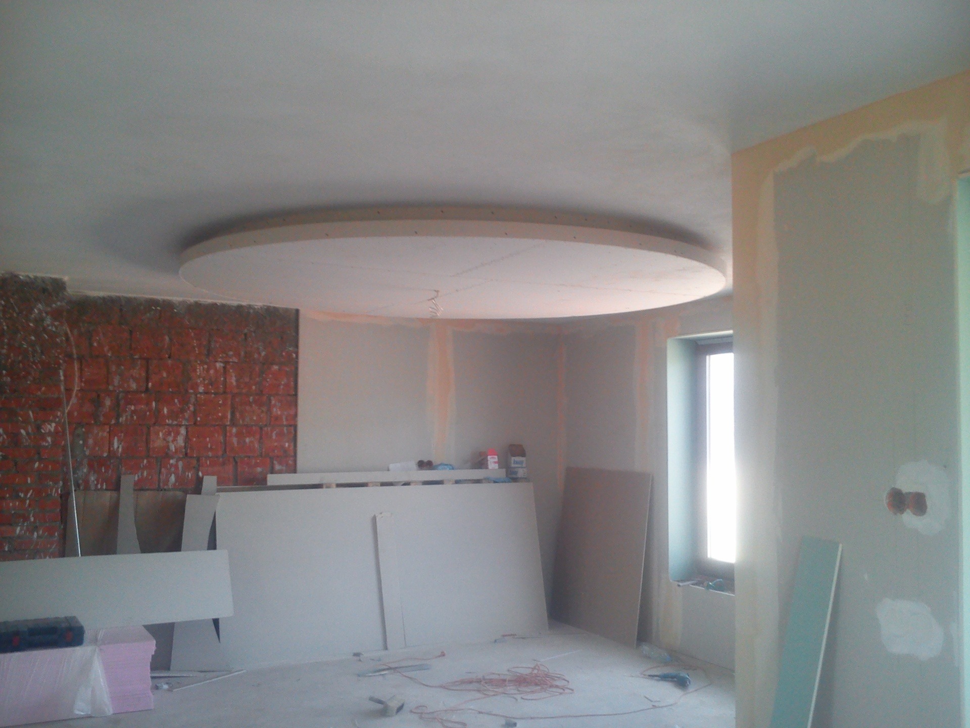 Wnętrze w trakcie remontu z okrągłym podwieszanym elementem sufitu, widoczne płyty gipsowe, fragment ściany z cegły oraz okno.