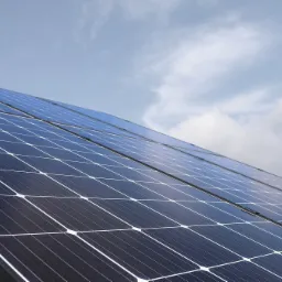 Rząd paneli słonecznych na dachu, widoczne mocowania, pochmurne niebo w tle.