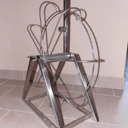 Metalowa konstrukcja o nietypowym, artystycznym kształcie, przypominająca szpulę z metalowymi prętami, na trójkątnej podstawie, stojąca na kafelkach.