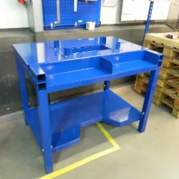 Niebieski, metalowy stół warsztatowy z półką, wykonany z profili zamkniętych, widoczny na tle szarej podłogi i niebieskiej ściany warsztatu.