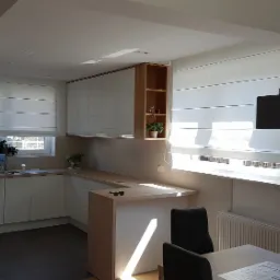 Jasna kuchnia z białymi szafkami, drewnianym blatem i roletami rzymskimi w oknach, widziana z perspektywy jadalni z ciemnymi krzesłami.