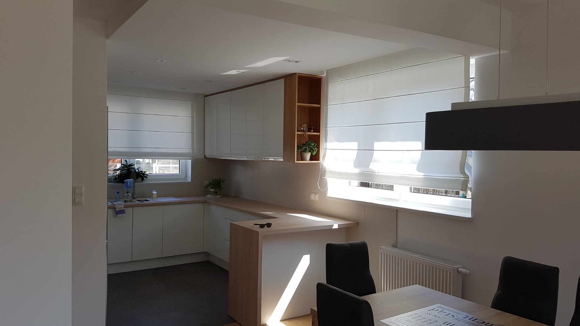 Jasna kuchnia z białymi szafkami, drewnianym blatem i roletami rzymskimi w oknach, widziana z perspektywy jadalni z ciemnymi krzesłami.