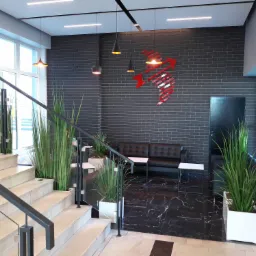 Nowoczesne wnętrze biurowe z marmurową podłogą, schodami z metalową balustradą, roślinami w białych donicach, szarą ceglaną ścianą z czerwonym logo, czarną sofą i stolikami.