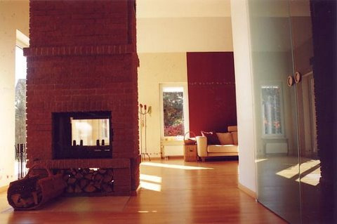 Salon z ceglanym kominkiem, drewnianą podłogą i beżową sofą w tle, oświetlony naturalnym światłem wpadającym przez okno.