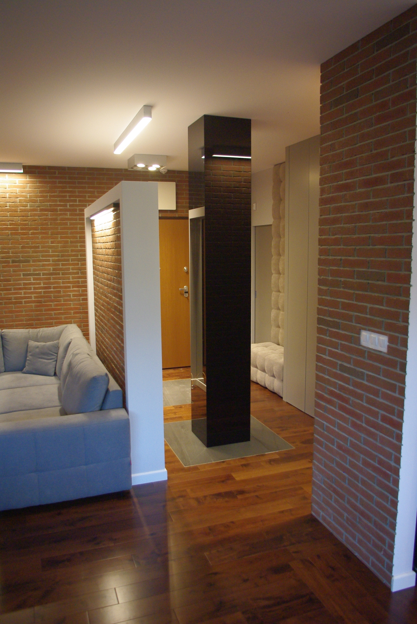 Nowoczesne wnętrze mieszkania z ceglaną ścianą, drewnianą podłogą, szarą sofą i czarnym, pionowym elementem dekoracyjnym z podświetleniem.