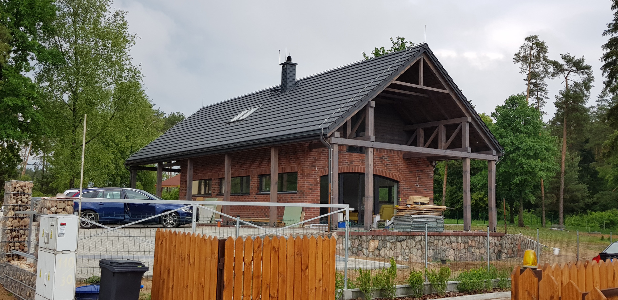 Nowoczesny dom jednorodzinny z czerwonej cegły z ciemną dachówką i drewnianą konstrukcją zadaszenia, widok zza drewnianego płotu.