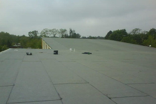 Widok z góry na płaski dach pokryty papą bitumiczną, z widocznymi elementami wentylacyjnymi i narzędziami pracy, tło stanowią drzewa i pochmurne niebo.