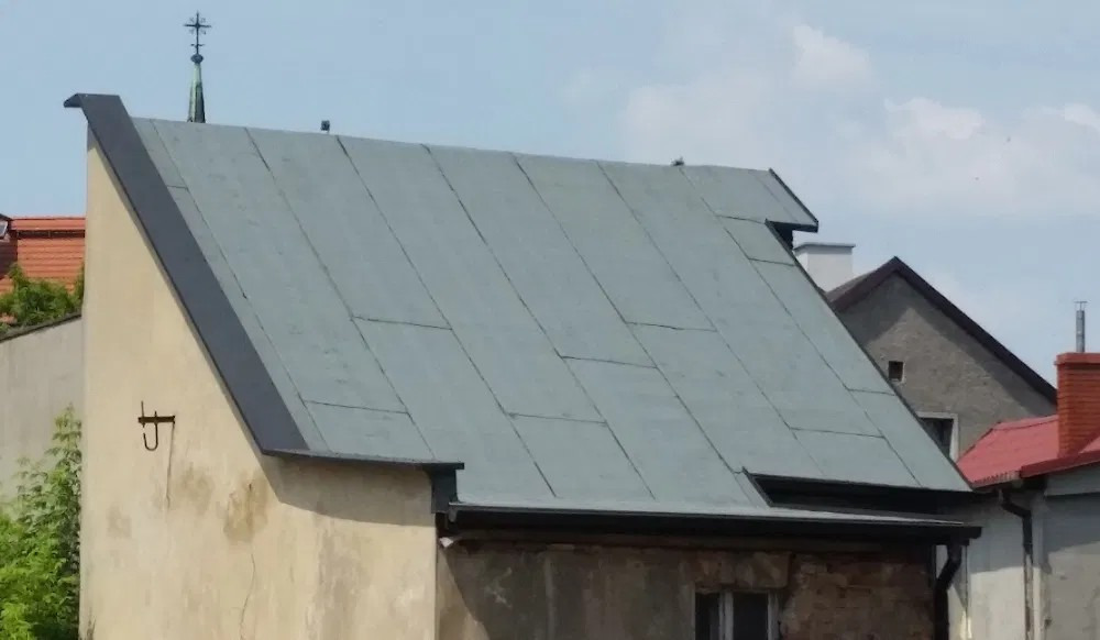 Szary dach z blachy trapezowej na starym budynku, widoczny hak na ścianie, w tle inne budynki i wieża kościelna.