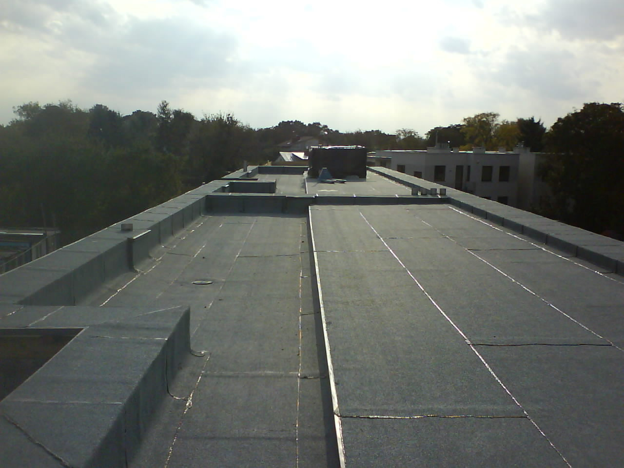 Płaski dach pokryty papą termozgrzewalną z widocznymi łączeniami i obróbkami blacharskimi, na tle budynków i drzew w pochmurny dzień.