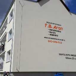 Ocieplanie elewacji bloku w Radziszewie, widoczni pracownicy na balkonie i przy ziemi podczas prac, na elewacji widnieje reklama firmy F.B. Aron z informacjami kontaktowymi.