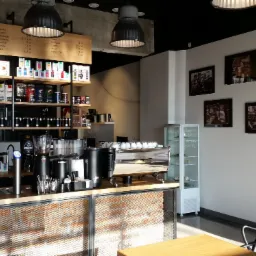 Wnętrze kawiarni z drewnianym blatem, eksponowanymi cegłami za siatką, półkami z kawami i napojami, oraz czarno-białymi zdjęciami na ścianie, oświetlone lampami w stylu industrialnym.