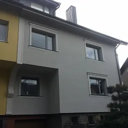 Dwukondygnacyjny dom z elewacją w odcieniach szarości, z dekoracyjnymi obramowaniami okiennymi i kontrastującą żółtą częścią sąsiedniego budynku.