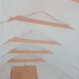 Poddasze z drewnianą konstrukcją pokryte białą pianką izolacyjną, widoczne fragmenty ścian z płyt OSB.