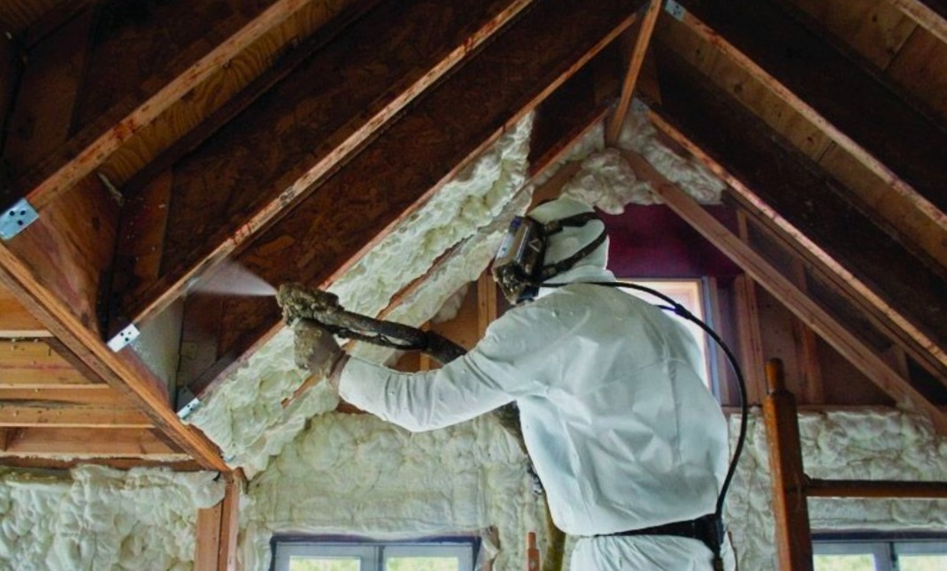 Pracownik w kombinezonie ochronnym aplikuje piankę izolacyjną na drewnianą konstrukcję poddasza, widoczne świeżo nałożone warstwy piany.