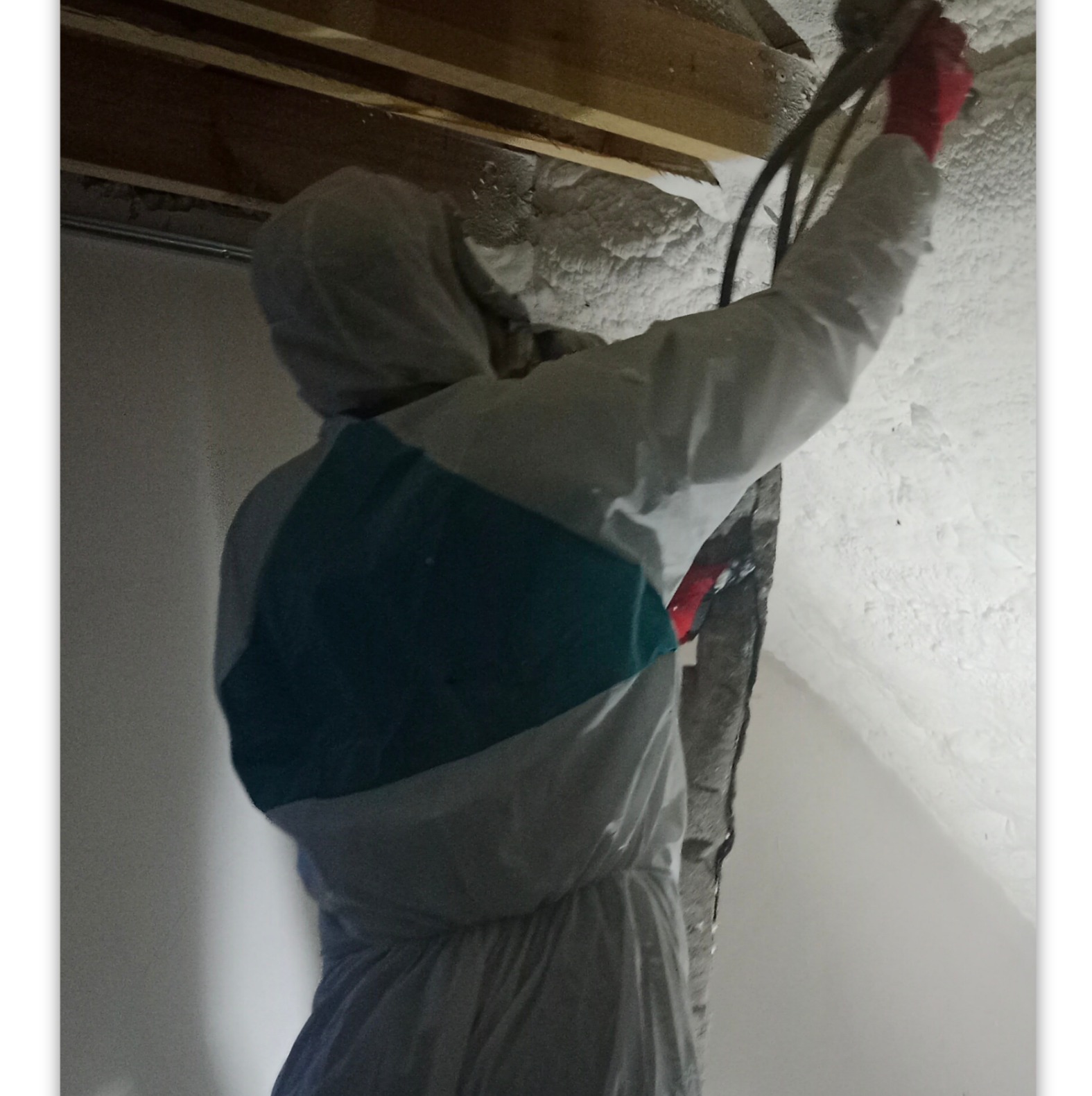 Pracownik w kombinezonie ochronnym aplikuje piankę izolacyjną na poddaszu, widoczne drewniane belki stropowe i częściowo pokryta pianą ściana.