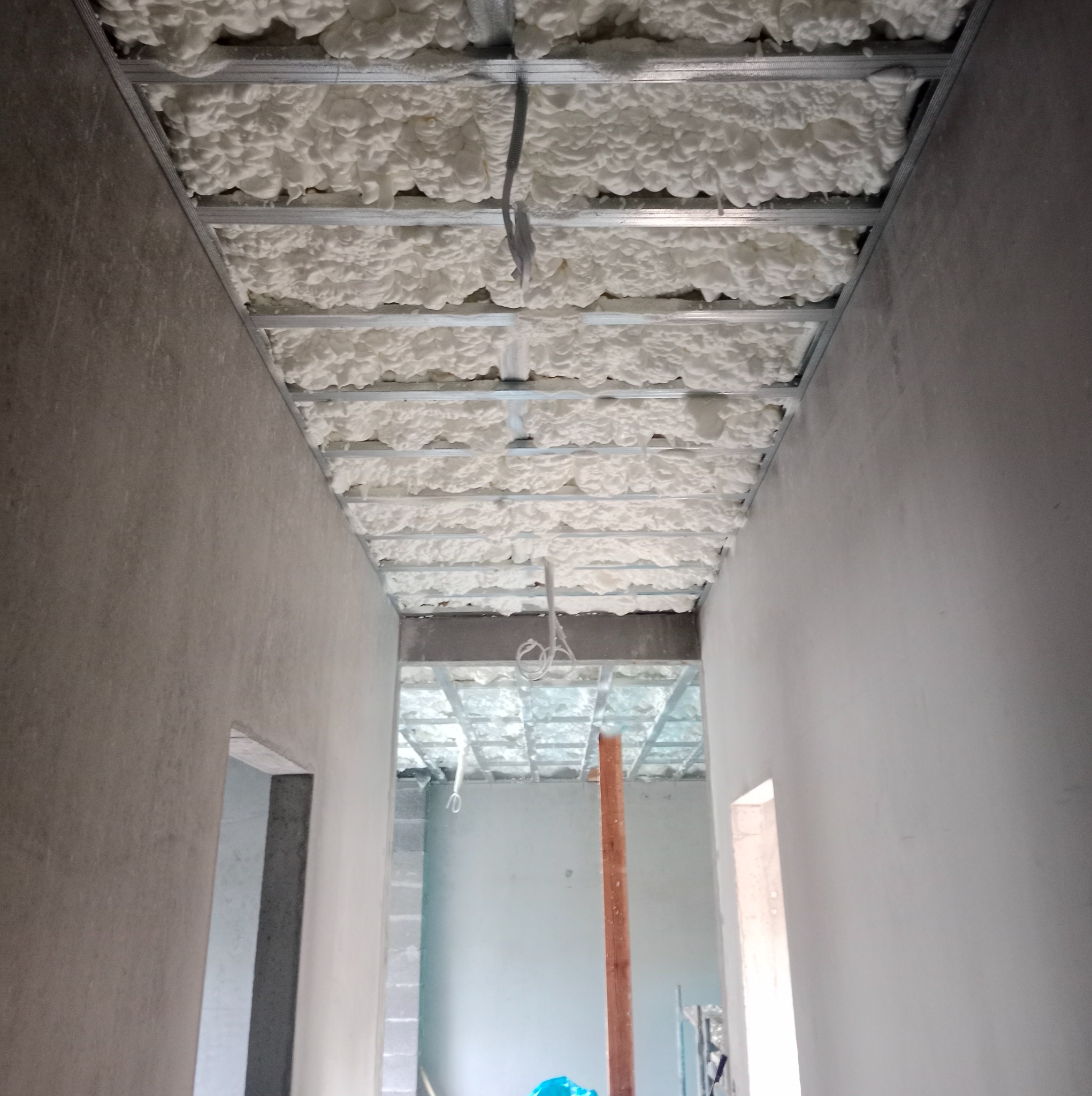 Poddasze w trakcie izolacji: biała pianka PUR wypełnia przestrzeń między krokwiami, widoczne metalowe profile konstrukcyjne, surowe ściany i otwory drzwiowe w tle.