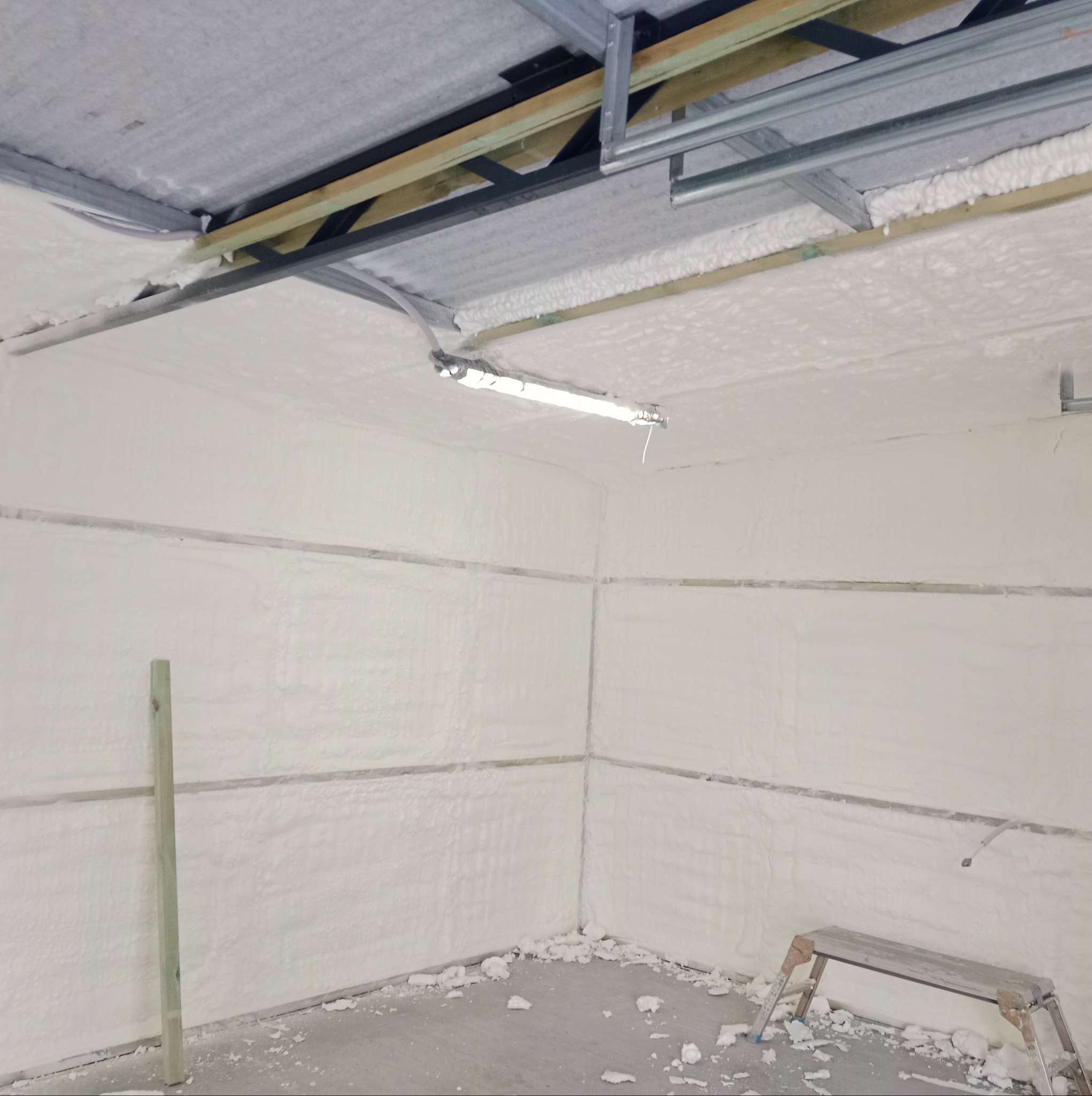 Wnętrze pomieszczenia po natrysku pianki poliuretanowej na ściany i sufit, widoczne profile konstrukcyjne pod sufitem, resztki pianki na podłodze i drabinka.