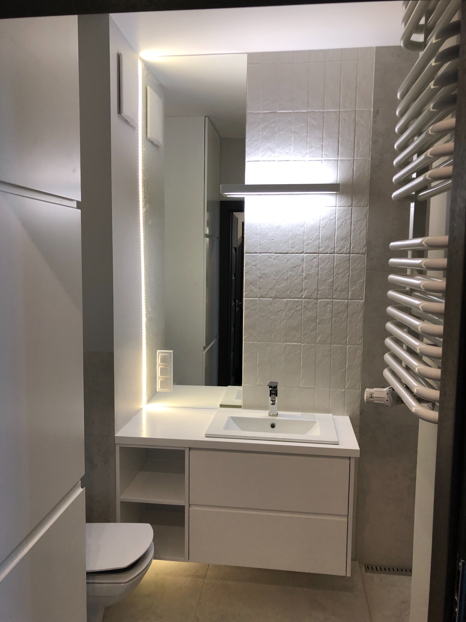 Nowoczesna łazienka w odcieniach szarości i bieli, z podświetlanym lustrem i grzejnikiem drabinkowym, widok na umywalkę z szafką i toaletę.