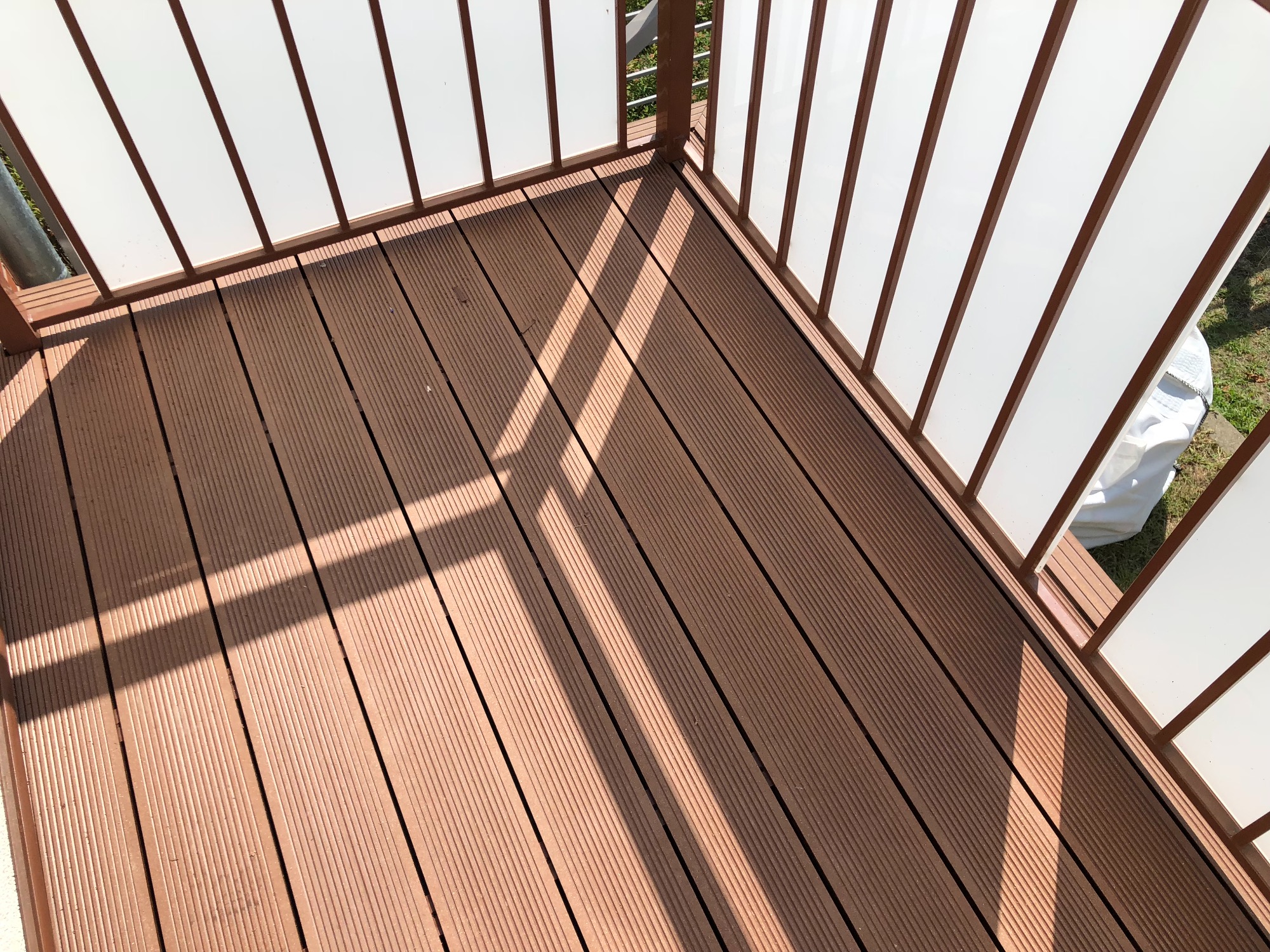 Nowoczesny balkon z brązową podłogą z desek kompozytowych i białymi panelami balustrady, oświetlony promieniami słońca tworzącymi geometryczne cienie.