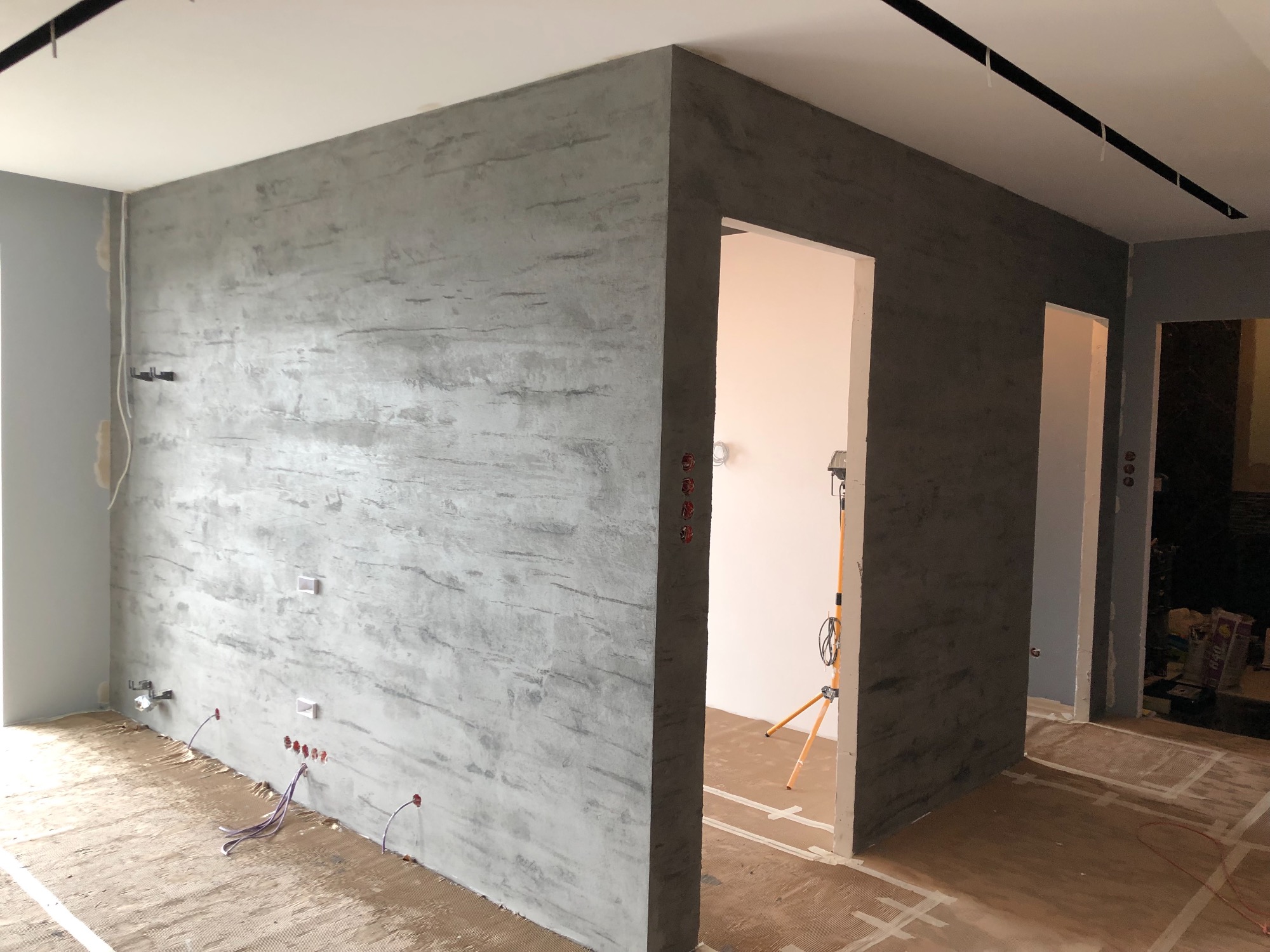 Surowa ściana z efektem betonu w trakcie remontu mieszkania, widoczne wystające kable elektryczne i przygotowana podłoga, oświetlenie robocze w przejściu.