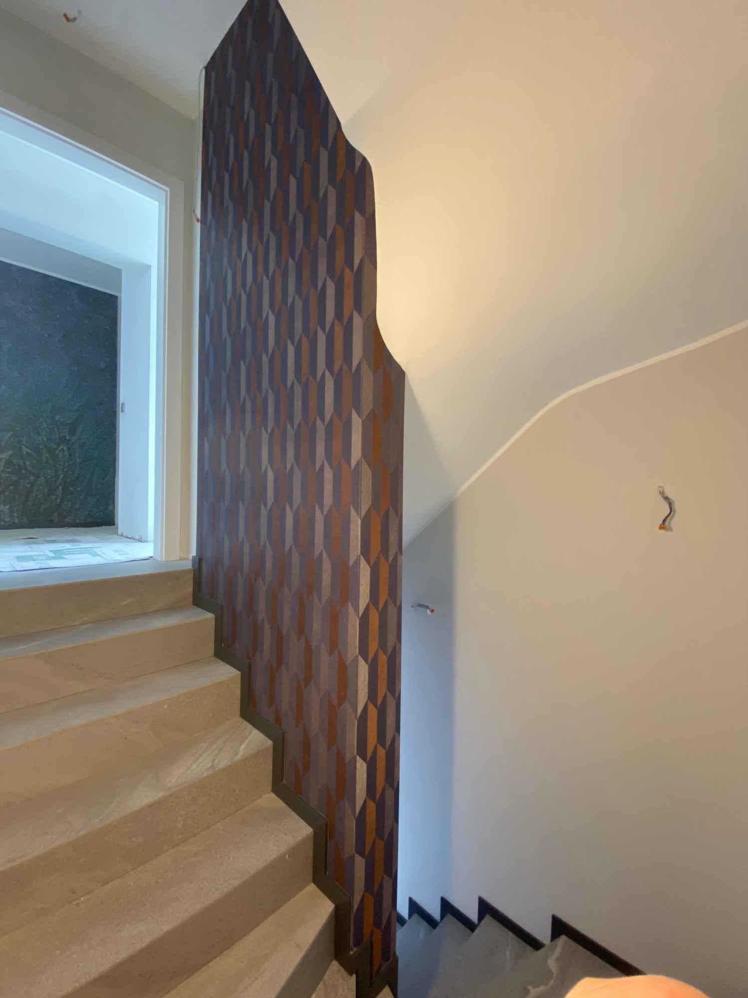 Klatka schodowa w trakcie remontu, widoczne schody wyłożone jasnym kamieniem, ściana z geometryczną tapetą w odcieniach brązu i szarości, oraz fragment instalacji elektrycznej wystający ze ściany.