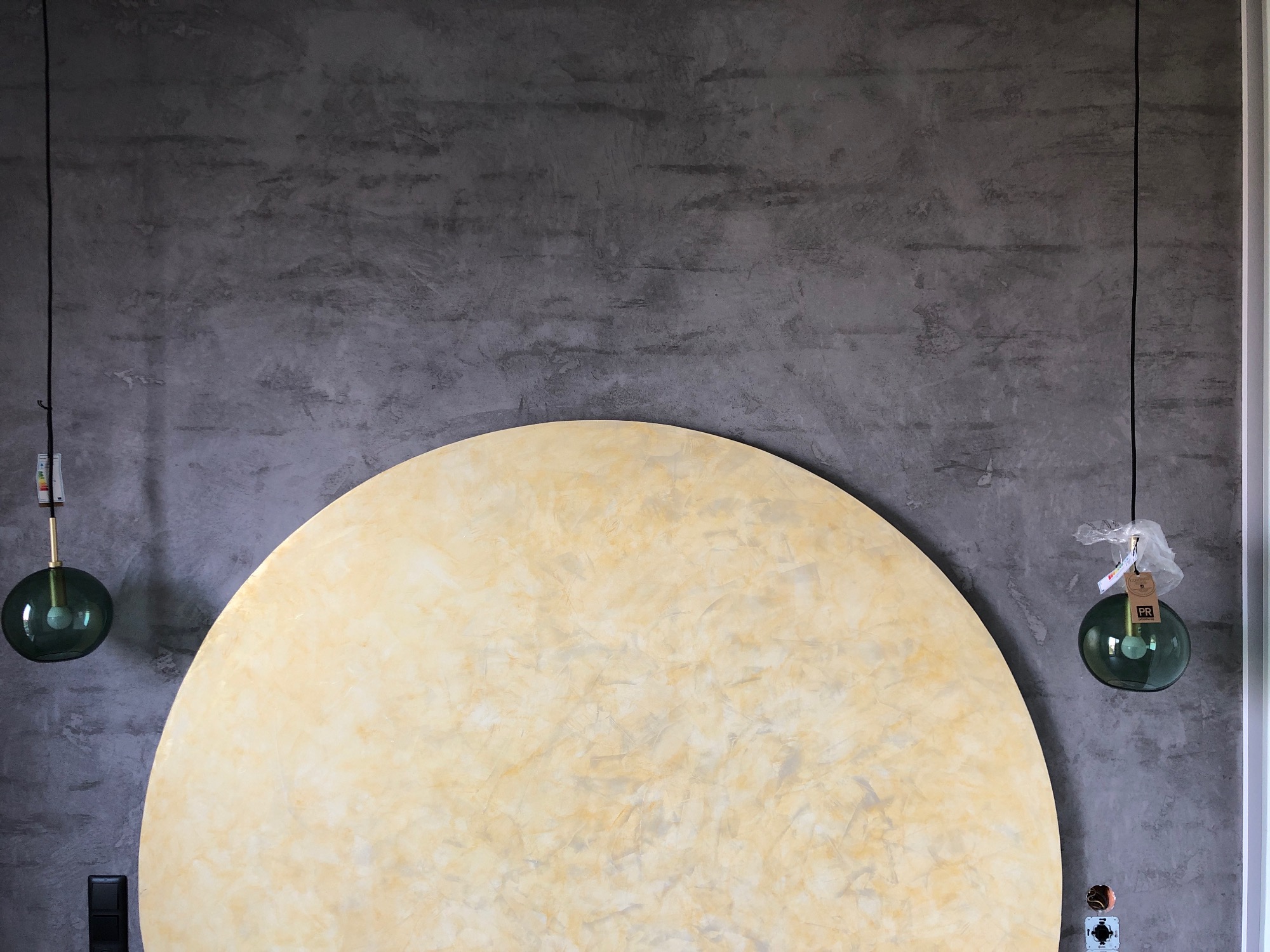 Dekoracyjna ściana z szarą fakturą betonu, okrągły element dekoracyjny w odcieniach beżu i wiszące szklane lampy w kolorze butelkowej zieleni, z widocznymi etykietami.