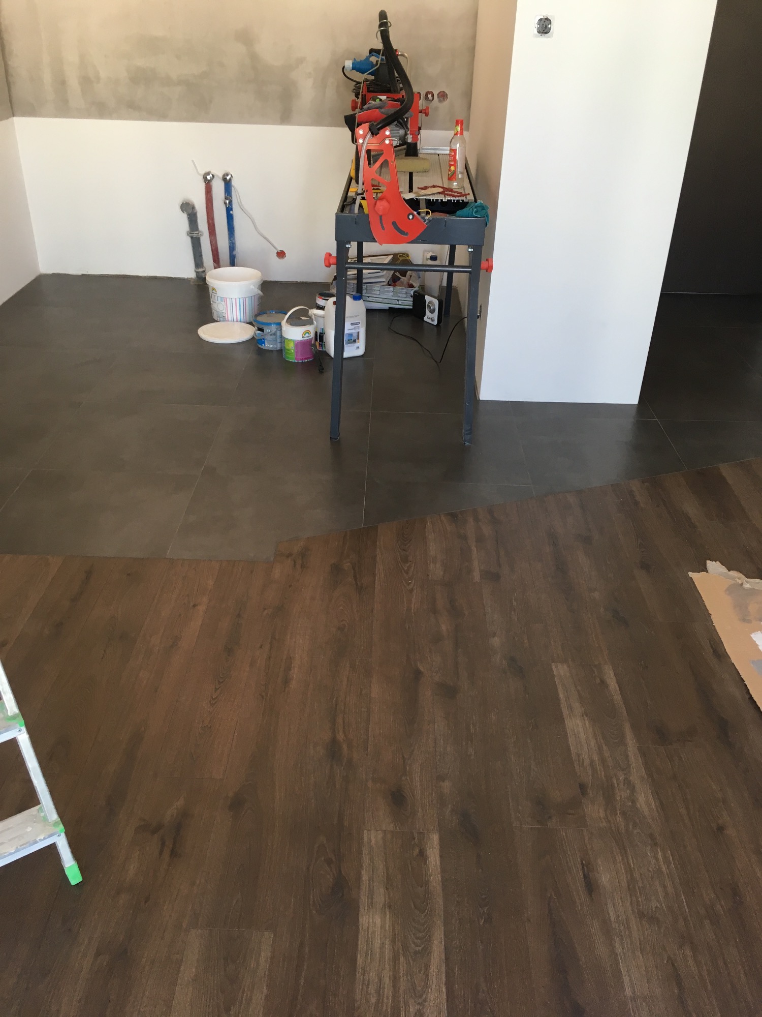 Proces łączenia ciemnych płytek ceramicznych z panelami podłogowymi w trakcie remontu, widoczne narzędzia na stole roboczym i fragment drabiny.