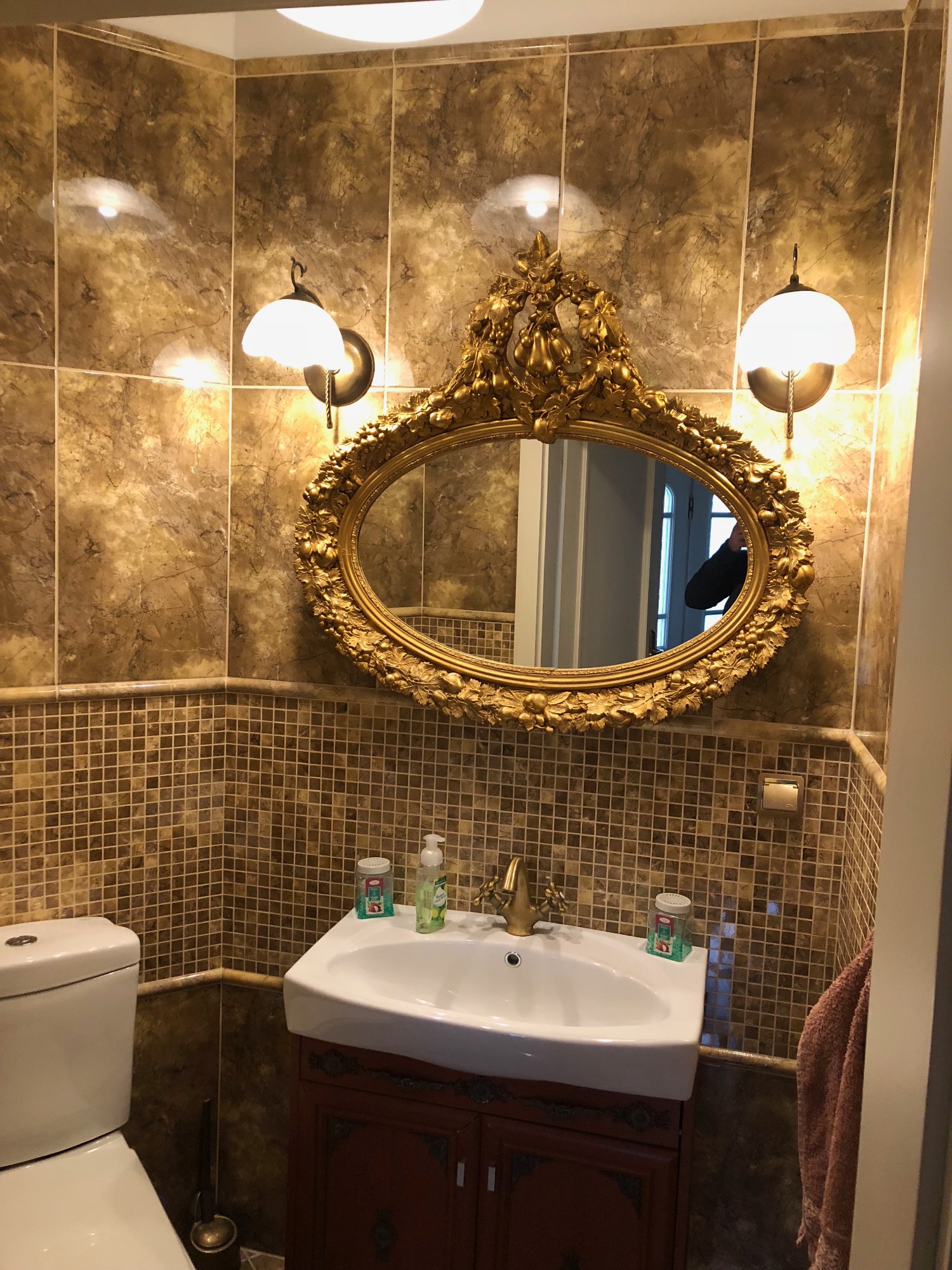 Wykończona łazienka w stylu retro z beżowymi płytkami, owalnym lustrem w złotej, zdobionej ramie, umywalką nablatową z brązową szafką i złotą baterią.