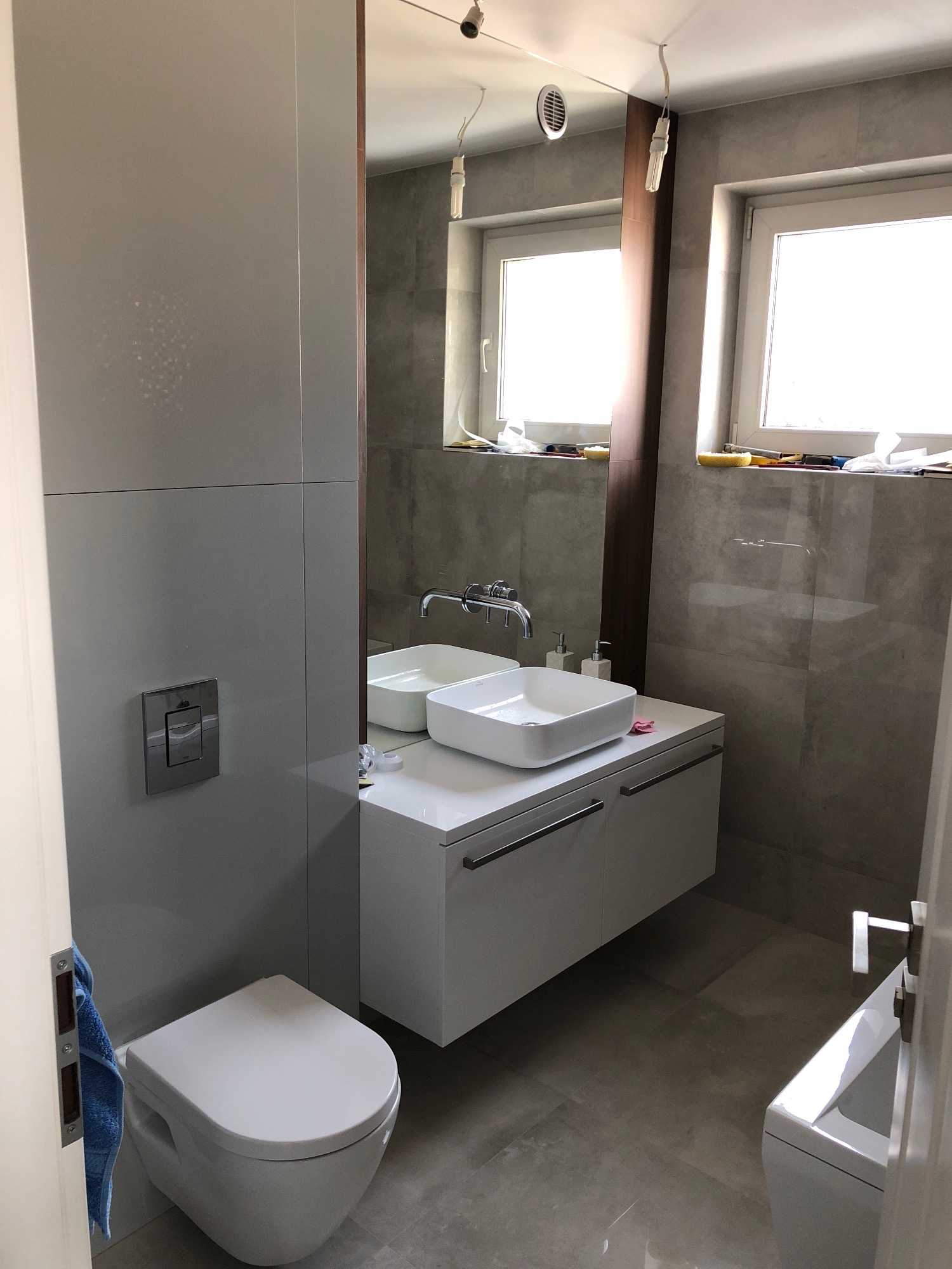 Nowoczesna łazienka z wiszącą toaletą, umywalką nablatową i szarą glazurą, widoczne okna z akcesoriami malarskimi na parapecie.