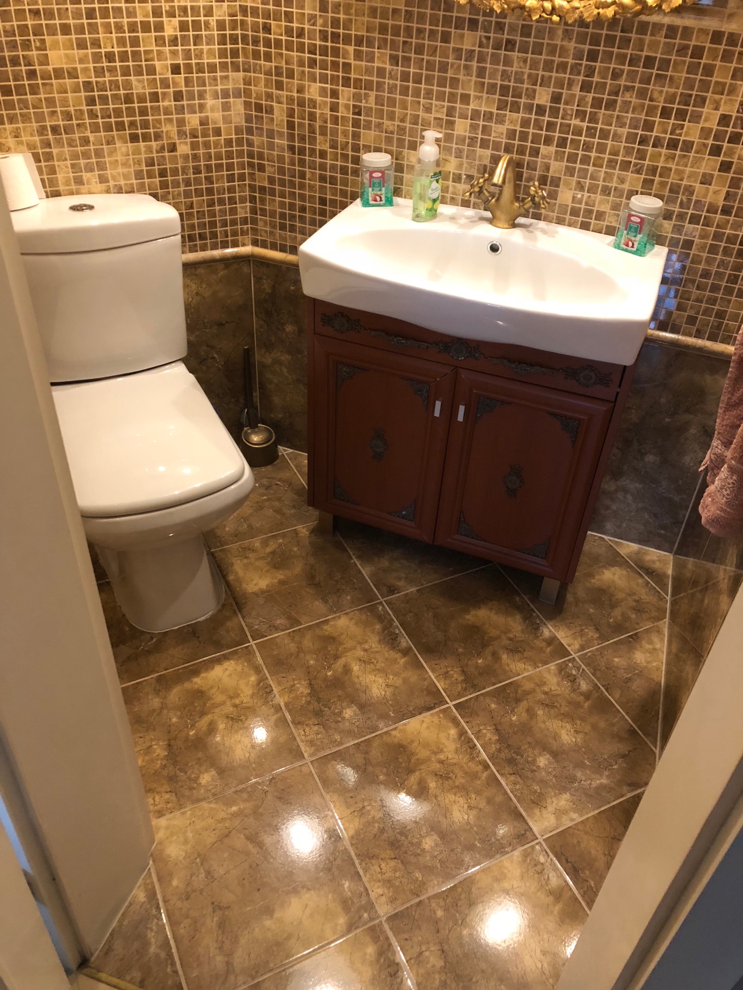 Ukończona łazienka z mozaikową ścianą, brązowymi płytkami podłogowymi, białą toaletą i umywalką na szafce w kolorze bordowym z ozdobnymi detalami.