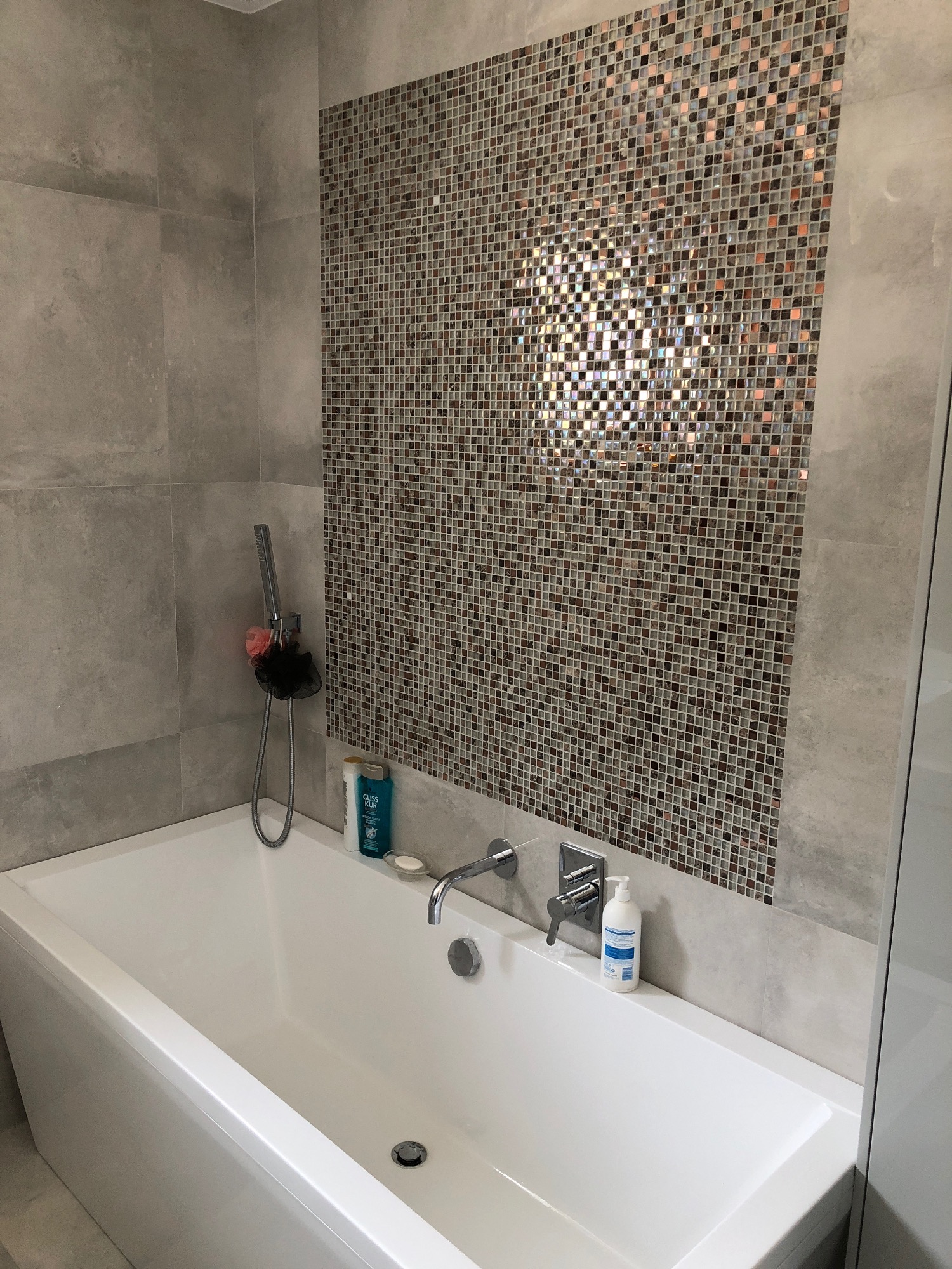 Nowoczesna łazienka z białą wanną, mozaiką na ścianie i szarymi płytkami, z widocznymi kosmetykami i słuchawką prysznicową.