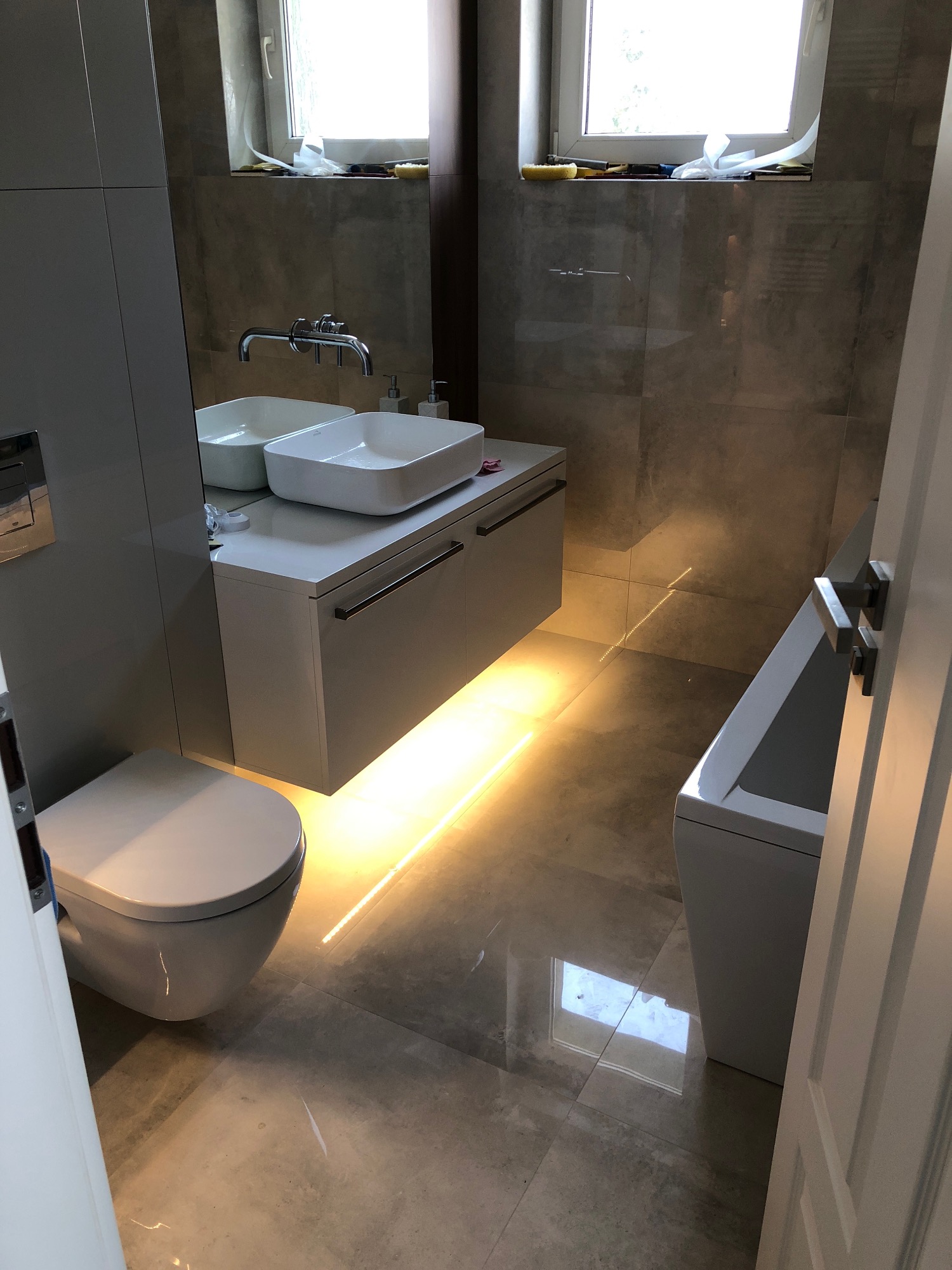 Nowoczesna łazienka z podświetlaną szafką pod umywalką, wiszącą toaletą i wanną, wykończona płytkami w odcieniach szarości.